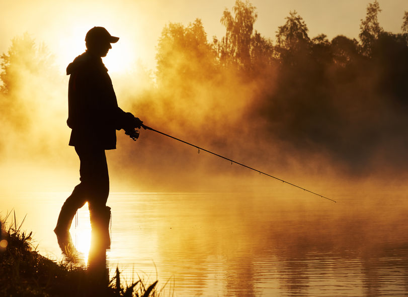 Fisher man fishing with spinning rod on a river bank at misty foggy sunrise